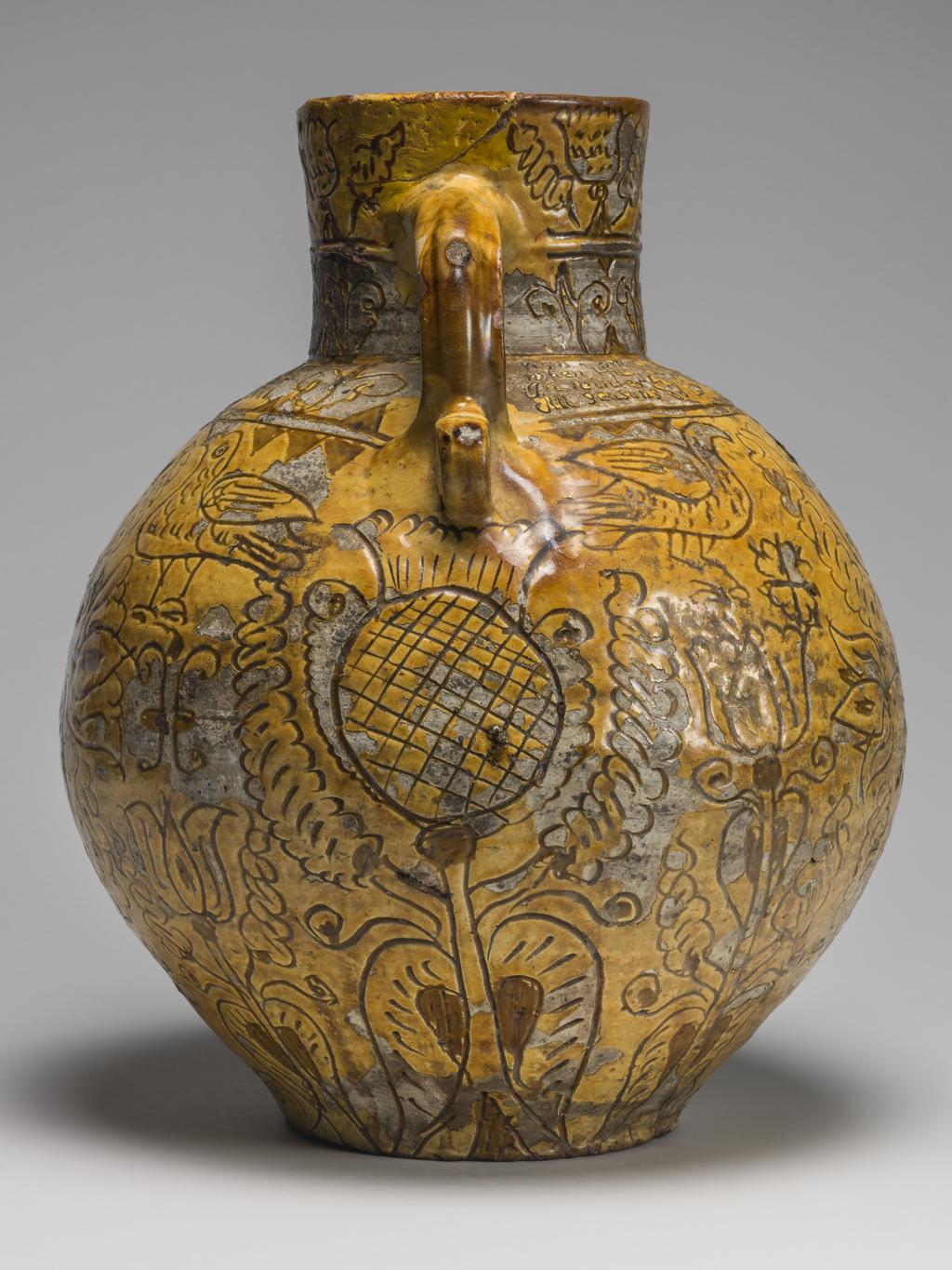 An image of Harvest Jug. Bulbous body, with a short neck pinched into a lip in front, and loop handle coiled up at lower end. On the front inscribed are the initials and date 'WH 1724', enclosed in a heart, with a thistle below and the crude busts of women, each above a cartouche with the date repeated, on either side. Below the handle is a thistle, whilst over the rest of the body except the shoulder are birds and formal tulips. On the shoulder is the inscription: 'Now i am come for to supply your workmen when in haruist dry when they do labour hard and sweat good drink is better far than meat. In winter time when it is cold i likewayes then good ale can hold All seasons do the same require and most men so strong drink desire'. Round the neck are two rows of flower motives. Earthenware, slip-coated with incised decoration under lead glaze, height, whole, 32.5 cm, dated 1724. English, North Devon.