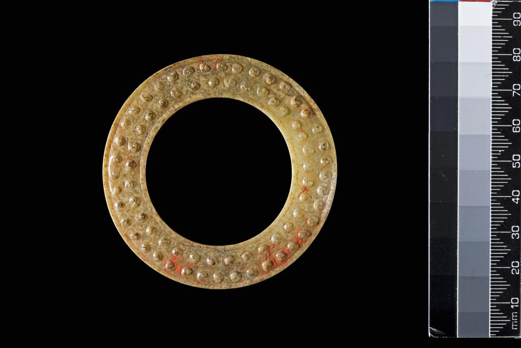An image of Ring. Grey white jade with small brown speckled areas, carved in the shape of a ring, decorated with raised spirals and the surface covered with a layer of cinnabar. It would have formed part of a pendant set. Nephrite, diameter 6.5 cm, 500-300 B.C. Chinese.