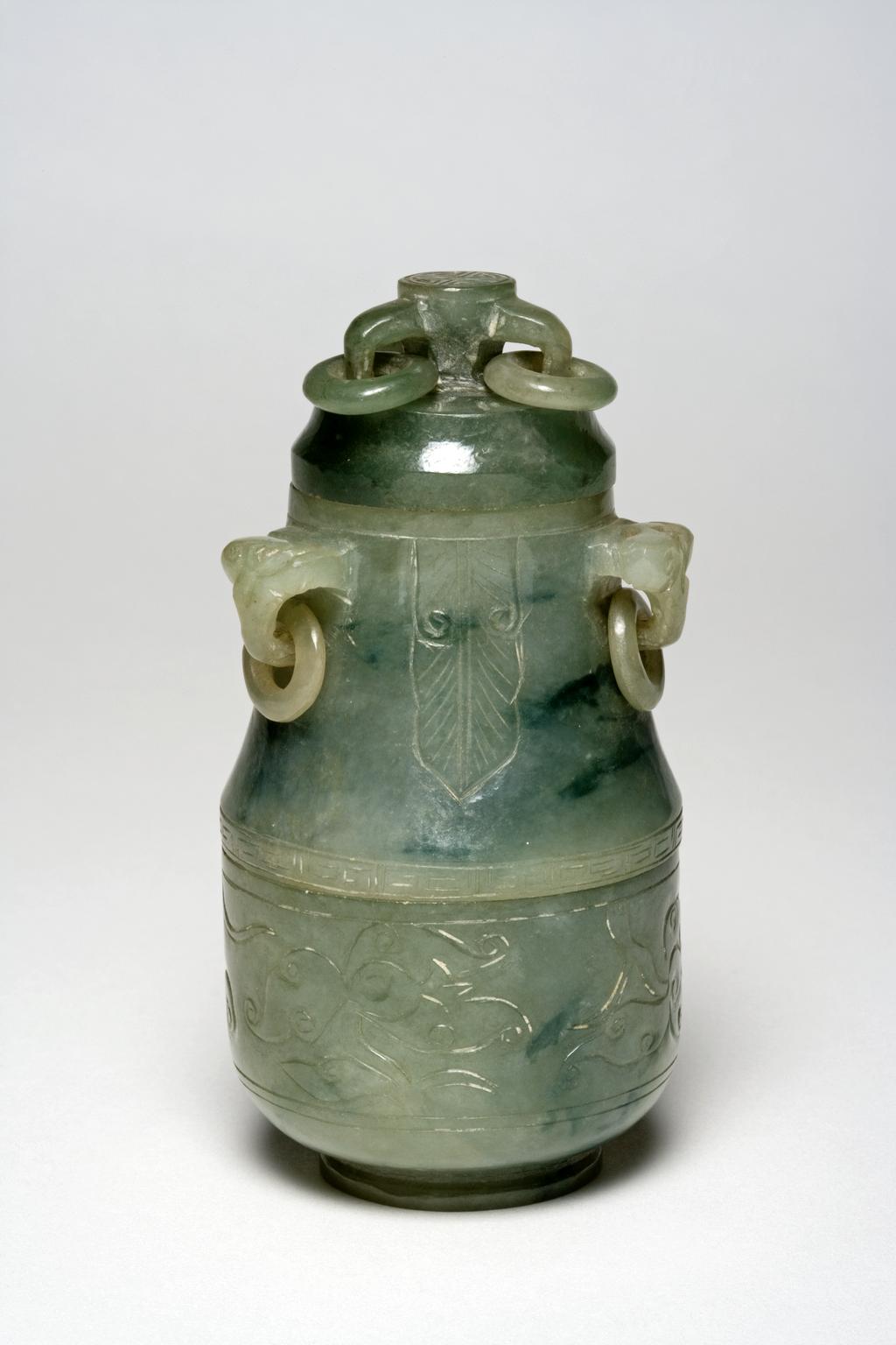 An image of Vessel/Double Box. Pale green jadeite with splashes of apple-green, carved as a double box and lid. The lid decorated with three loose rings, the main body with three banana leaves between three loose ring handles, and the lower part with floral motifs. Jadeite, height 10.5 cm, 1800-1900. Chinese.