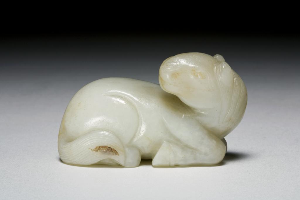 An image of Greyish green jade with light brown veining. The horse recumbent with its neck slightly arched as it looks back towards its tail. Its eyes are cocked up attentively and are shown in incised lines. The legs are folded beneath the body. The mane and the tail are strongly striated. Nephrite, length 6.65 cm, 1600-1800. Chinese.