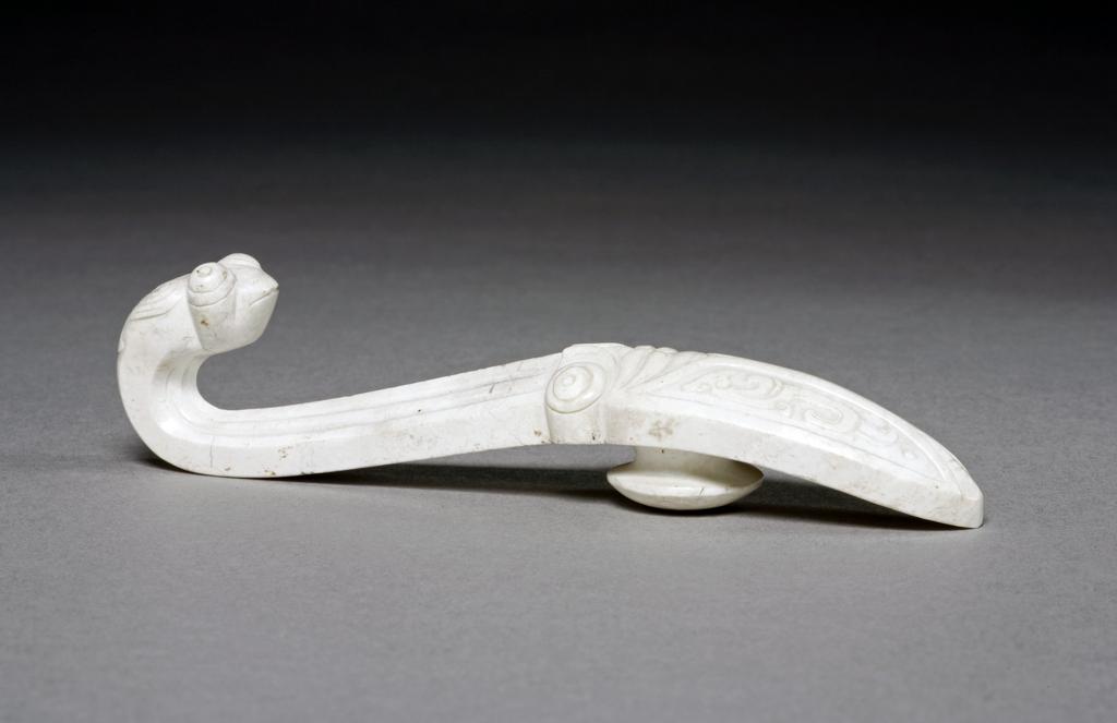 An image of Belt hook/dai gou. Opaque white jade with grey line inclusions. The body decorated with a cicada with a phoenix motif on its wings. The hook terminates with a praying mantis with prominent eyes. The topic of this carving refers to the well-known proverb: When the praying mantis is (intent on) catching the cicada, it fails to notice that the yellow bird is behind (waiting to catch the praying mantis). Nephrite, length 11.1 cm,1600-1800, Chinese.