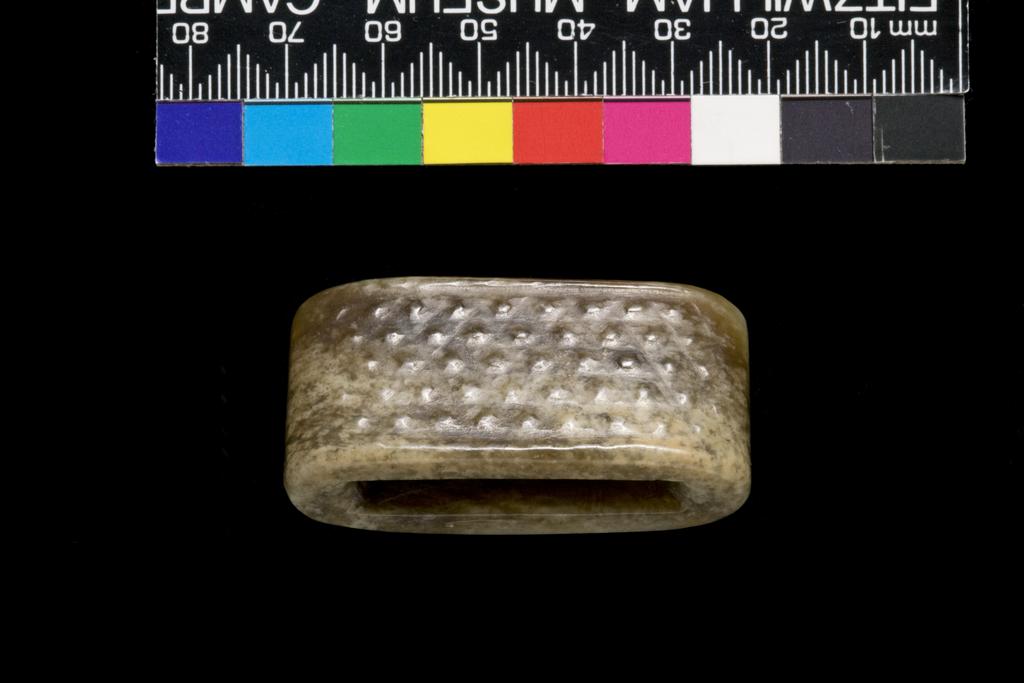 An image of Scabbard Slide. Grey-green jade with large areas of brown staining. One side decorated with C-shaped volutes and the other with rows of small spirals in relief. Nephrite, height 1.45 cm, length 4.6 cm, width 2.2 cm, 1368-1644 AD, Chinese.