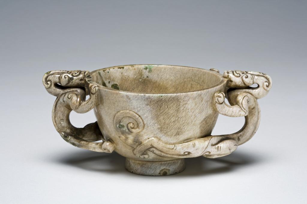 An image of Cup/ Greyish jade cup resting on a tall footring. The handles are decorated with feline dragons in high relief. Incised on the footring the character yong, eternal. Jade cups in this form are very commonly seen in the late Ming dynasty. It is possible that this object has been treated to give the appearance of having been buried and of great age. Nephrite, height 6 cm, width 14.7 cm, 1500-1700. Chinese.
