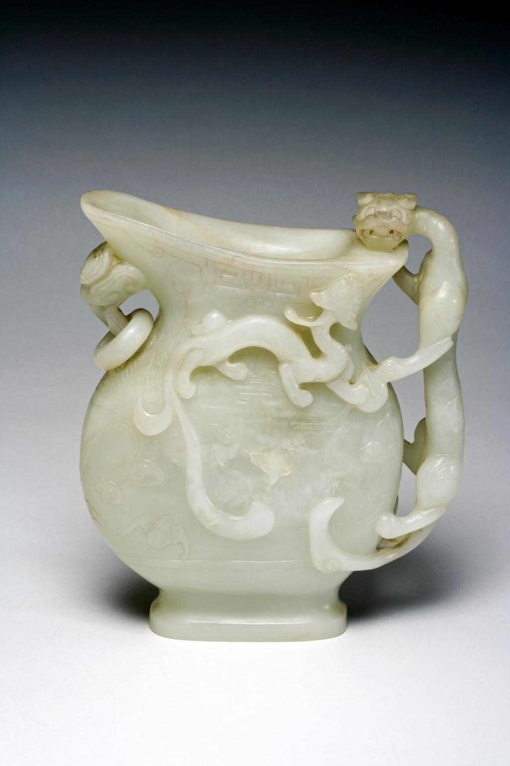 An image of Pale green jade jug, carved in high relief with a feline dragon clambering over the rim and forming the handle. Beneath the rounded spout is a handle decorated with lingzhi fungus, from which is suspended a loose ring. On both sides the main surfaces are decorated with clouds, three bats and an archaic motif from ancient bronzes. Nephrite, height, 16.4 cm, width, 14.8 cm, 1800-1900. Chinese.