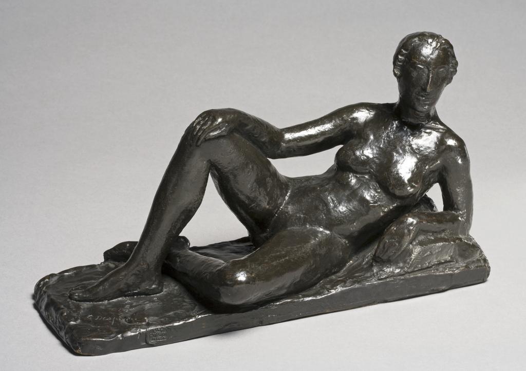 An image of DESPIAU, Charles'Reclining Female Nude'Bronze, cast, with dark brown patina, slightly polished.. The reclining figure rests on her left elbow; the right knee is raised and the right hand and arm rest on it; the left leg, flat on the ground, is crossed under the other. The left elbow is raised on a pillow. The figure is attached to its base.FranceC.1922