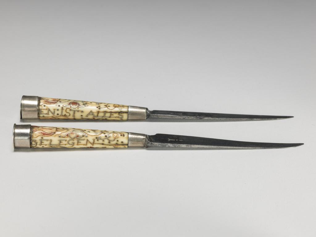 An image of Cutlery/Pair of knives and case. Unknown handle maker and case maker. Unidentified cutler. Object probably either German or English. Ivory, silver, enamel and steel, shagreen and white metal; each knife has a steel tapering blade and ivory handle of circular section with silver ferrule and terminal cap. Both ivory handles are inscribed and decorated with scrolls and tulips in inlaid silver wires and red enamel . Knife A is inscribed 'AN: GOTTES: SE / EN: IST: ALLES' and knife B is inscribed 'GELEGEN / ANNO: 1674'. The case (C) is of wood covered with unpolished pearl ray shagreen with a white metal mount to the tip. It has a pull-off lid, is of lobed oval section and tapers to a point. Length, knife A, 13.5 cm, length, knife B, 13.3 cm, length, case, 17.3 cm, circa 1674. English.