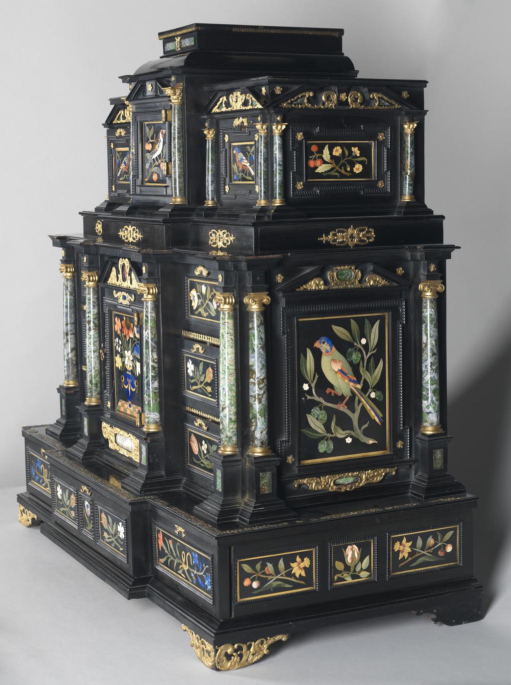An image of Augsburg 17th century silver-gilt mounted ebony Table Cabinet with Florentine pietre dure plaques from the Grand Ducal workshops. Boscher, Elias (1600-1674). Of architectural form, the stepped superstructure fitted with three frieze drawers above a central cupboard door, architectural pediment and inset with a panel depicting a chaffinch on a fruiting bough, flanked by columns of Smaragdite Gabbro with Corinthian capitals flanked by two deep drawers, inset with panels depicting parrots within fruiting boughs, with three further fitted drawers below, the large central cupboard door inset with a panel of a lapis lazuli flower filled urn issuing stems including a carnation, anemone, daffodil and lilies and enclosing an architectural interior, flanked by further Smaragdite Gabbro Ionic columns and three drawers to each side inset with further symmetrical floral panels including tulips, roses, carnations, lilies and bluebells, on shaped bracket feet, each side inset with large central panels of parrots on fruiting boughs, with four further floral panels above and below, the reverse veneered in kingwood, amaranth and stained walnut in geometric stellar designs. The cabinet containing a complex series of fourty-four secret drawers and compartments to the interior. The cabinet is signed in pencil on the underside 'Elias Boscher' and 'gemacht'. The central cabochon mount stamped with the Augsburg town mark and the silversmiths mark of Johann Spitzmacher (active 1655-1678). The front bears the ebony stamp "EBEN" with the pinecone of Augsburg. Height 85 cm, width 84cm, depth 40cm, circa 1660.