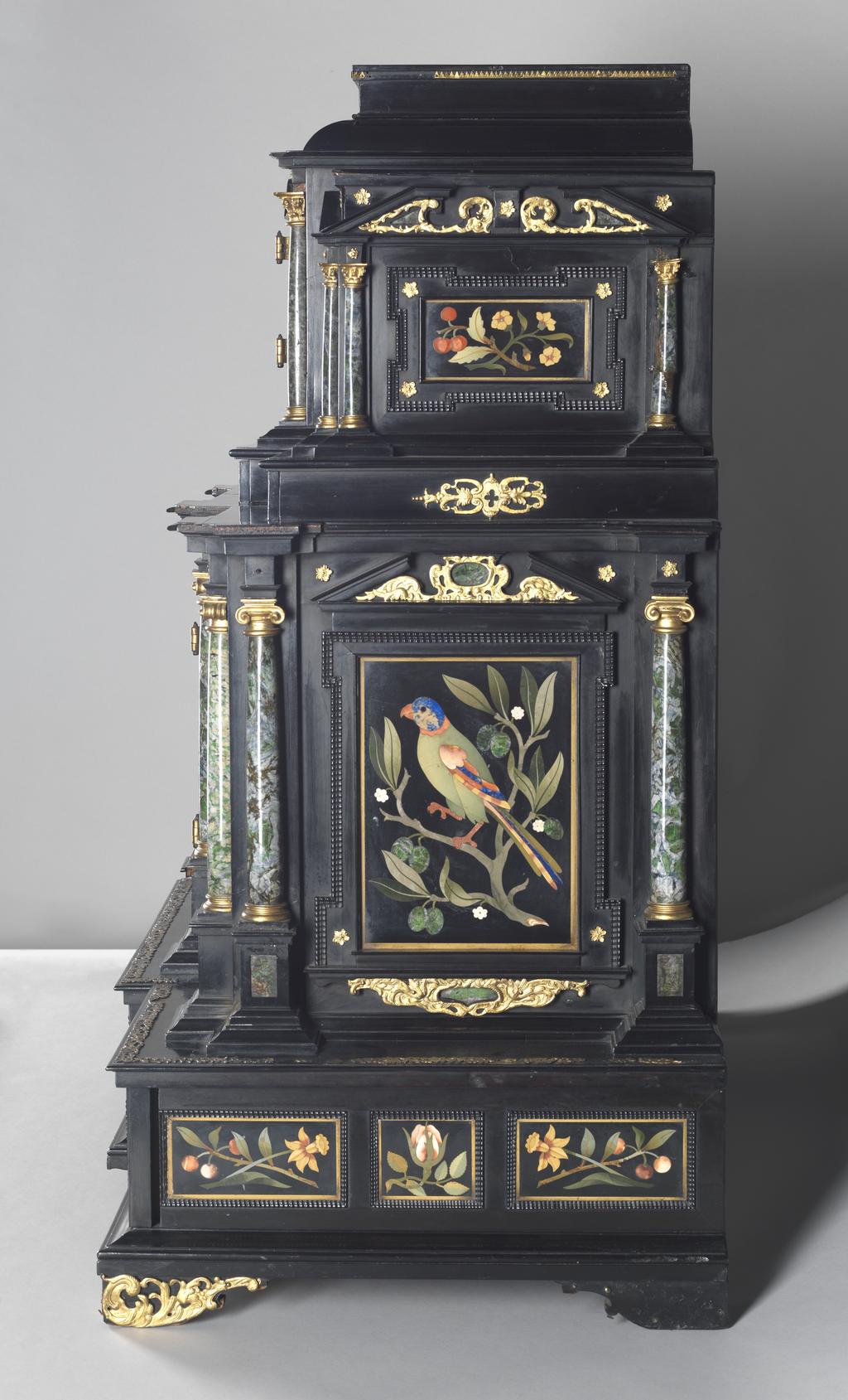 An image of Augsburg 17th century silver-gilt mounted ebony Table Cabinet with Florentine pietre dure plaques from the Grand Ducal workshops. Boscher, Elias (1600-1674). Of architectural form, the stepped superstructure fitted with three frieze drawers above a central cupboard door, architectural pediment and inset with a panel depicting a chaffinch on a fruiting bough, flanked by columns of Smaragdite Gabbro with Corinthian capitals flanked by two deep drawers, inset with panels depicting parrots within fruiting boughs, with three further fitted drawers below, the large central cupboard door inset with a panel of a lapis lazuli flower filled urn issuing stems including a carnation, anemone, daffodil and lilies and enclosing an architectural interior, flanked by further Smaragdite Gabbro Ionic columns and three drawers to each side inset with further symmetrical floral panels including tulips, roses, carnations, lilies and bluebells, on shaped bracket feet, each side inset with large central panels of parrots on fruiting boughs, with four further floral panels above and below, the reverse veneered in kingwood, amaranth and stained walnut in geometric stellar designs. The cabinet containing a complex series of fourty-four secret drawers and compartments to the interior. The cabinet is signed in pencil on the underside 'Elias Boscher' and 'gemacht'. The central cabochon mount stamped with the Augsburg town mark and the silversmiths mark of Johann Spitzmacher (active 1655-1678). The front bears the ebony stamp "EBEN" with the pinecone of Augsburg. Height 85 cm, width 84cm, depth 40cm, circa 1660.