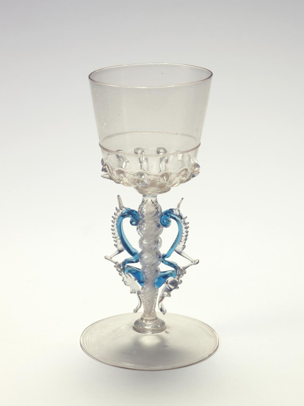 An image of Wine Glass. Unknown production, probably Venice. Approximately a bucket-shaped bowl with ribbed base. A double fillet at the mid-way point. Stem with fine small knops, spinal ribbing and tapering towards a plain foot. Blue wings with colourless denticulations. Crizzling. Lead-glass, blown bowl, ribbed lower part of bowl and stem spine, height, whole, 6 1/4 in, diameter, whole, 3 1/4 in, circa 1600-1650. Façon de Venise. Venetian.