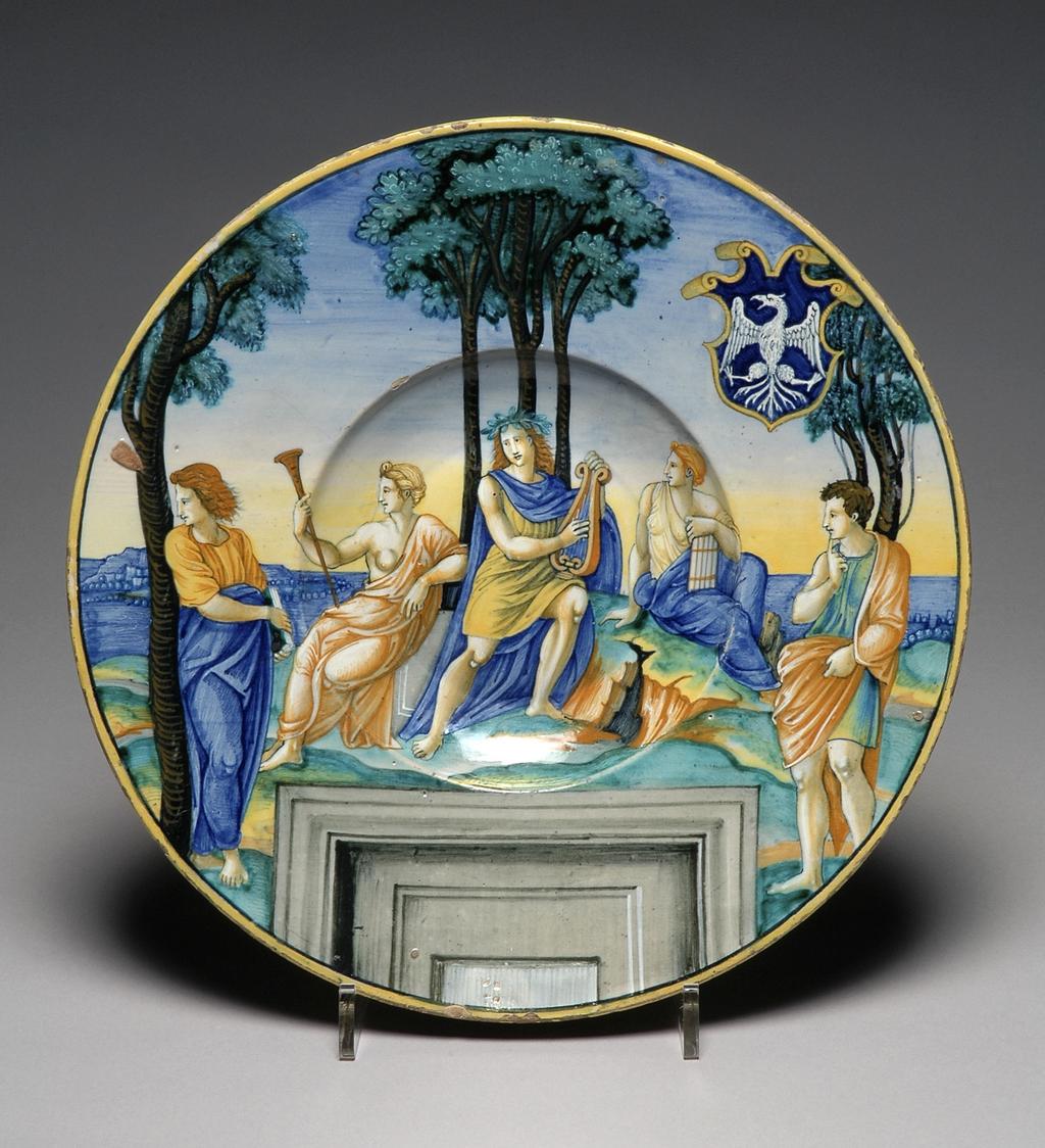 An image of Maiolica dish, painted in polychrome with Mount Parnassus, Apollo, Euterpe and Erato. Milan Marsyas Painter, probably, Italy, The Marches, Urbino. Shape 55. Circular with broad, sloping rim, shallow well and convex centre. Parnassus. In the middle, Apollo sits playing a lyre in front of three trees. On the left, the Muse Euterpe sits holding a trumpet, and on the right, the Muse Erato, holding pipes. On each side a poet stands beside a tree; the one on the left looks away from the central group, the other towards it, holding his right index finger to his lips. Behind the figures there is a landscape and below, a window frame. On the upper right side there is a shield of Tuscan form, charged with the arms azure, an eagle displayed argent. The edge is yellow. Pale yellowish buff earthenware, tin-glazed overall. There is a fault in the glaze on the left of the rim and on the reverse it is pale beige and thinly applied. Painted in blue, green, yellow, orange, brown, black, grey, and white. Height, whole, 4.1 cm, diameter, whole, 30.7 cm, circa 1530-1531. Renaissance. Notes: The scene was derived from the engraving by Marcantonio Raimondi after a preparatory drawing by Raphael for his fresco of Parnassus, in the Stanza della Segnatura in the Vatican. All the figures other than Apollo, two Muses and two poets have been omitted; the trees and, curiously, the window frame, have been retained, and a landscape background has been inserted.