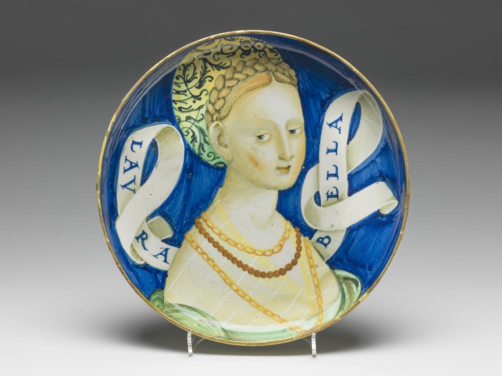 An image of Maiolica. Shallow bowl on low foot. Unidentified potter. This type of dish is traditionally attributed to Castel Durante, but could have been made in Urbino. A bust of a young woman, three-quarters to right, her gaze cast downwards to her right. Her blonde hair is dressed in a plait over the crown of her head. She wears a green and yellow turban embroidered with black scrolling foliage; two gold chains and a rope of beads; and a green dress with a yellow chemise showing on the shoulders. A scroll inscribed `LAVRA/BELLA' in blue projects on either side of her head. The background is blue. On the back are four concentric yellow bands: on the edge of the foot, just beyond the junction of the foot and bowl, and two round the rim. Pale buff earthenware, tin-glazed overall. Painted in blue, green, yellow, orange, cream, and black. Height, whole, 4.7 cm, diameter, whole, 22.7 cm, circa 1525-1535. Renaissance.