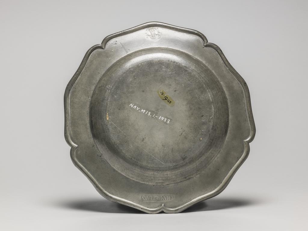An image of Plate. Thomas Chamberlain of King Street, Soho. Pewter, cast and hammered, with engraved arms, height, whole, 2 cm, diameter, whole, 25 cm, circa 1750-1775. Circular with five indentations and five outward curves, curved sides and flat base. The underside of the rim has an applied raised edge. The rim is engraved with tan almost circular shield bearing he unregistered coat-of-arms of David Garrick (1717-79), surrounded and foliage. This plate was part of a service made for the celebrated actor and manager of the Drury Lane Theatre, David Garrick (1717-79). If the arms had tinctures (colours) they would be described: 'Per pale or and azure on the dexter compartment a tower gules and on the sinister on a mount vert a sea horse azure mane, fins and tail of the first, on a chief gold, three mullets of the second. (See Documentation, Burke) Thirty-one pieces from the service bearing either Garrick's arms or crest are at the Shakespeare Birthplace Trust at Stratford-on-Avon. A.F. de Navarro Bequest.