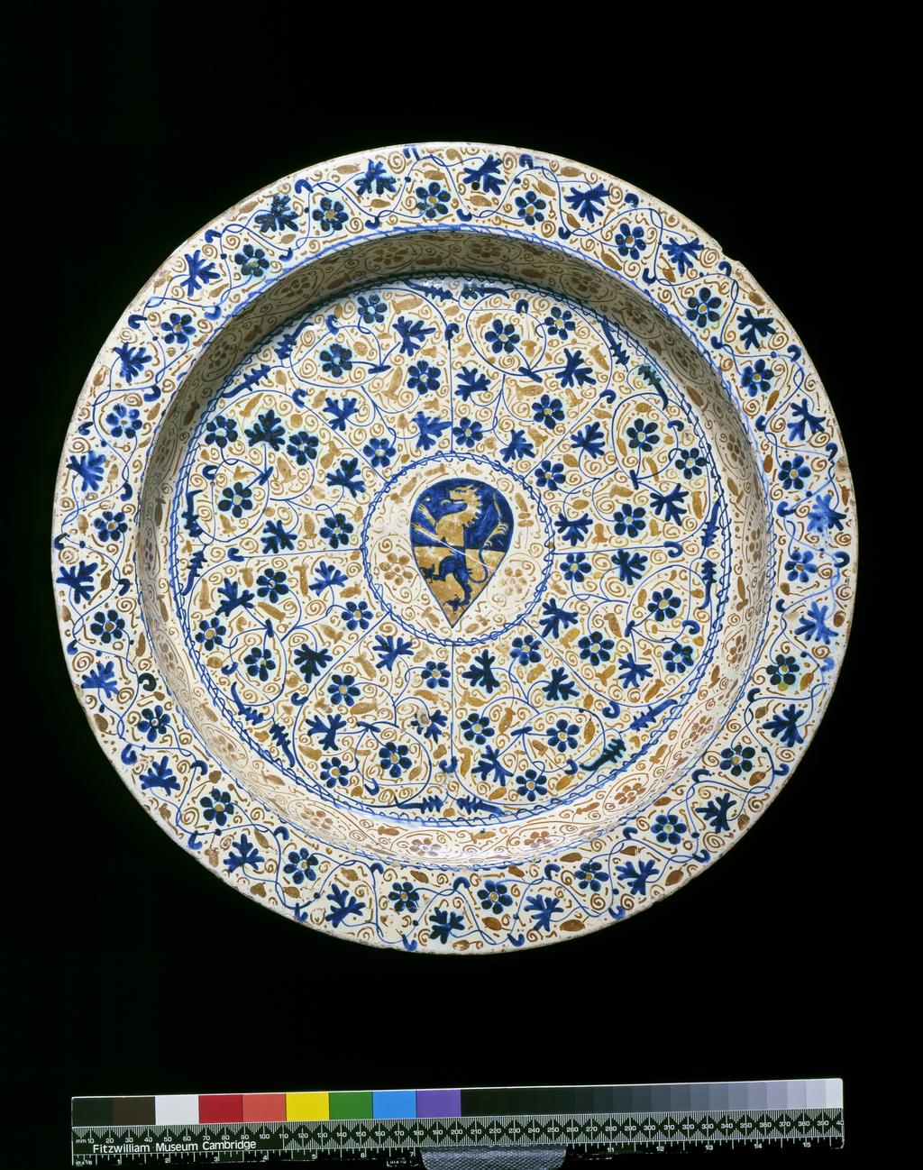 An image of Dish, BasinTin-glazed earthenware painted in cobalt-blue and copper lustreEarthenware, tin-glazed, and painted in cobalt-blue and copper lustre. Circular with flat rim, vertical sides, and flat central area. In the centre there is a heater-shaped shield bearing the arms per fesse azure and or a lion rampant countercharged within a blue spirally-entwined circle, from which eight curling sprays of bryony flowers and parsley-like leaves radiate towards another spirally entwined circle. The sides are decorated in lustre only, and the rim in blue and lustre with scrolling and wavying sprays of similar flowers and leaves. The back is decorated in lustre only with a central six-petalled flower surrounded by circlets and dotsSpanishC.1450-75