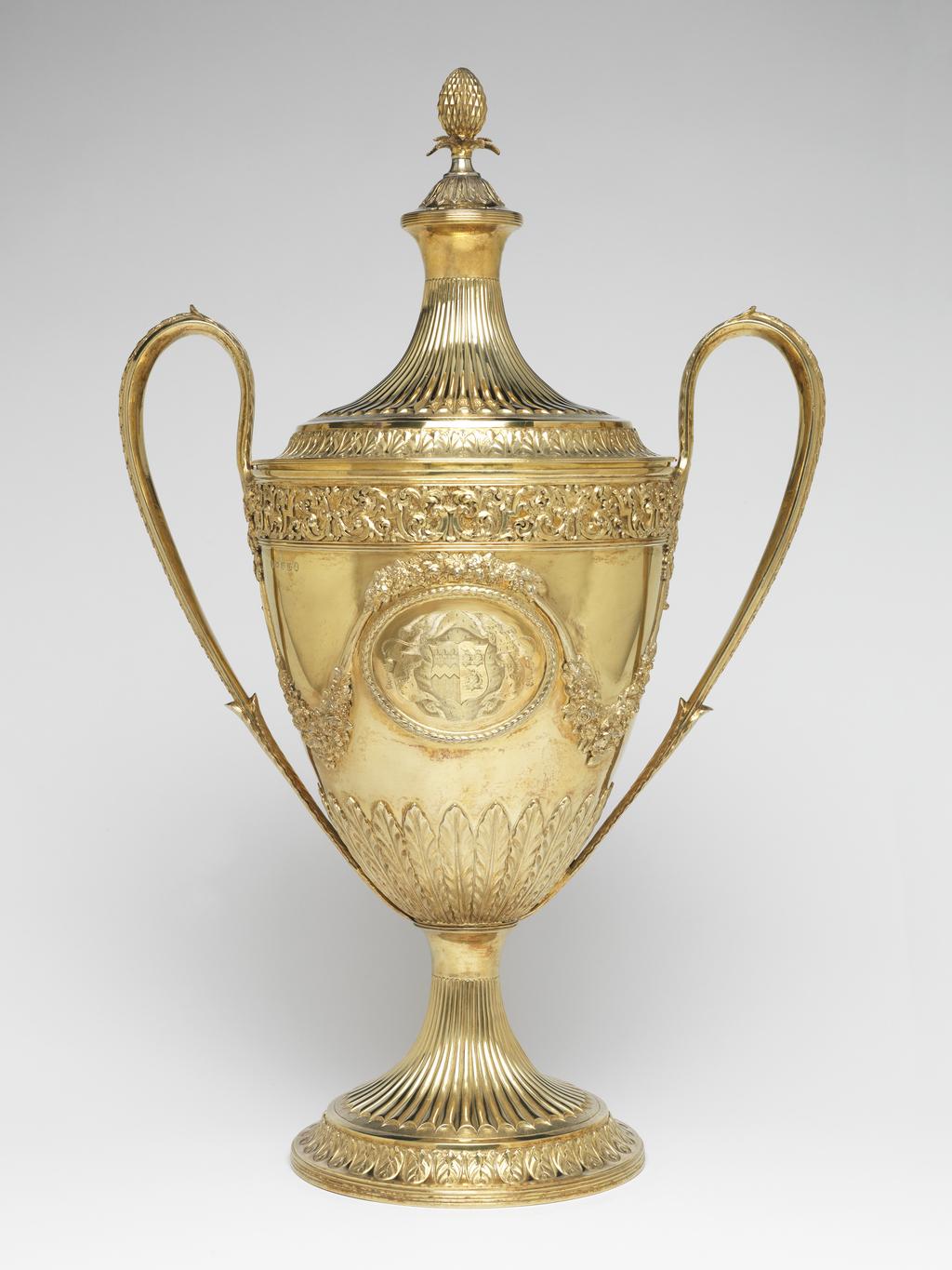 An image of Race Cup. Bateman, Peter. Bateman, Ann. Production Place:  London. The circular foot has a reeded edge above which is a band of leaf and bud ornament on a matted ground; the stem is embossed with bat's wing inverted fluting with an area of matting at the lower end of the flutes. The lower part of the tapering bowl has a calyx of lanceolate, wavy-edged leaves, above which, on each side of the body, is an oval medallion framed by husks. One medallion contains a plaque, cast and chased with two race horses and their jockeys racing in a landscape; the other is engraved with a coat of arms, sable a fess dancetty or in chief three fleur de lis argent impaling azure three uniorns' heads erased argent 2 and 1. Above each medallion there is an applied festoon of flowers and foliage from which depend two more festoons on either side, which loop up to meet the handles. The upper rim of the bowl has an applied frieze of strapwork and scrolling folliage within two moulded borders. The handles have lanceolate wavy-edged leaves at each upper end and a rib of husks on the exterior. The decoration of the cover follows that of the foot, but rises to a dome with reeded border topped by wavy edged leaves and an inverted pineapple finial. Decorated with cast and chased, applied, embossed and matted ornament and engraved with a coat of arms. Silver-gilt, height, overall, 59.9 cm, width, overall, 38.7 cm, 1799-1800. Neoclassical. Notes: The coat-of-arms on the dexter side are for Feeke, Feake or Feke of Wighton, Norfolk, or Hewster of Norfolk. The arms on the sinister side have not been identified, but could be for Shelley of Norfolk if the field was gules not azure.