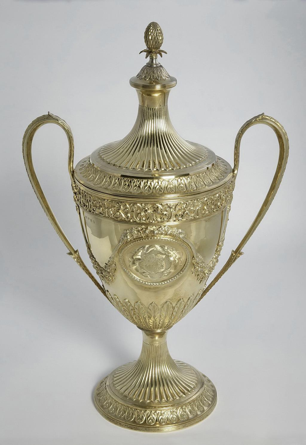 An image of Race Cup. Bateman, Peter. Bateman, Ann. Production Place:  London. The circular foot has a reeded edge above which is a band of leaf and bud ornament on a matted ground; the stem is embossed with bat's wing inverted fluting with an area of matting at the lower end of the flutes. The lower part of the tapering bowl has a calyx of lanceolate, wavy-edged leaves, above which, on each side of the body, is an oval medallion framed by husks. One medallion contains a plaque, cast and chased with two race horses and their jockeys racing in a landscape; the other is engraved with a coat of arms, sable a fess dancetty or in chief three fleur de lis argent impaling azure three uniorns' heads erased argent 2 and 1. Above each medallion there is an applied festoon of flowers and foliage from which depend two more festoons on either side, which loop up to meet the handles. The upper rim of the bowl has an applied frieze of strapwork and scrolling folliage within two moulded borders. The handles have lanceolate wavy-edged leaves at each upper end and a rib of husks on the exterior. The decoration of the cover follows that of the foot, but rises to a dome with reeded border topped by wavy edged leaves and an inverted pineapple finial. Decorated with cast and chased, applied, embossed and matted ornament and engraved with a coat of arms. Silver-gilt, height, overall, 59.9 cm, width, overall, 38.7 cm, 1799-1800. Neoclassical. Notes: The coat-of-arms on the dexter side are for Feeke, Feake or Feke of Wighton, Norfolk, or Hewster of Norfolk. The arms on the sinister side have not been identified, but could be for Shelley of Norfolk if the field was gules not azure.