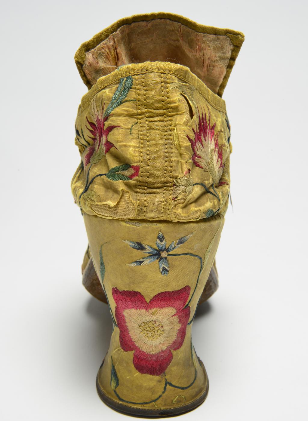 An image of Textiles. Shoes/pumps. Silk taffeta, bound with ribbon; floss silk embroidery in French knots, long and short, satin and split stitches; tied with ribbon. Ground covered by a pattern of trailing and scrolling stems with leaves and flowers, carnation, rose, etc., so arranged that there is a carnation on each toe and a rose on each heel with a five petalled blue flower above it. The left shoe has a rose, the right a carnation at each side of the seam. Silk, length, whole, 7 in, height, back, 5.25 in, height, heel, 3 in, circa 1700-circa 1749. English.