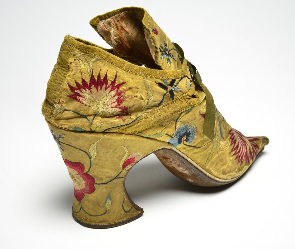 An image of Textiles. Shoes/pumps. Silk taffeta, bound with ribbon; floss silk embroidery in French knots, long and short, satin and split stitches; tied with ribbon. Ground covered by a pattern of trailing and scrolling stems with leaves and flowers, carnation, rose, etc., so arranged that there is a carnation on each toe and a rose on each heel with a five petalled blue flower above it. The left shoe has a rose, the right a carnation at each side of the seam. Silk, length, whole, 7 in, height, back, 5.25 in, height, heel, 3 in, circa 1700-circa 1749. English.