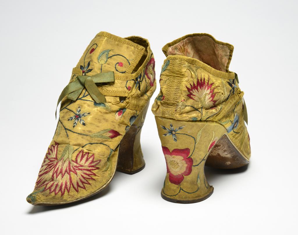 An image of Textiles. Shoes/pumps. Silk taffeta, bound with ribbon; floss silk embroidery in French knots, long and short, satin and split stitches; tied with ribbon. Ground covered by a pattern of trailing and scrolling stems with leaves and flowers, carnation, rose, etc., so arranged that there is a carnation on each toe and a rose on each heel with a five petalled blue flower above it. The left shoe has a rose, the right a carnation at each side of the seam. Silk, length, whole, 7 in, height, back, 5.25 in, height, heel, 3 in, circa 1700-circa 1749. English.