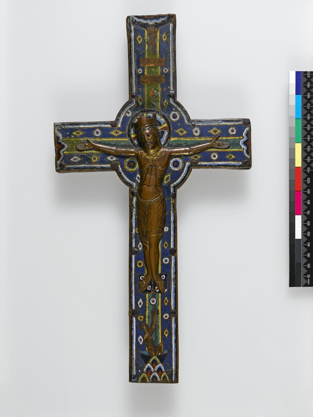 An image of Crucifix, with an applied bronze figure of Christ. Ground of dark blue is relieved with lozenges and circles of many colours. The centre of the cross is dark green shading off to a yellow. At the foot are the rocks of Golgotha and a beardless figures rising from a tomb. Oak, covered with copper, champlevé and enamelled, height 34 cm, width 18 cm, depth 1.5 cm, circa 1200 to 1300. Limoges, France. Romanesque, Medieval.