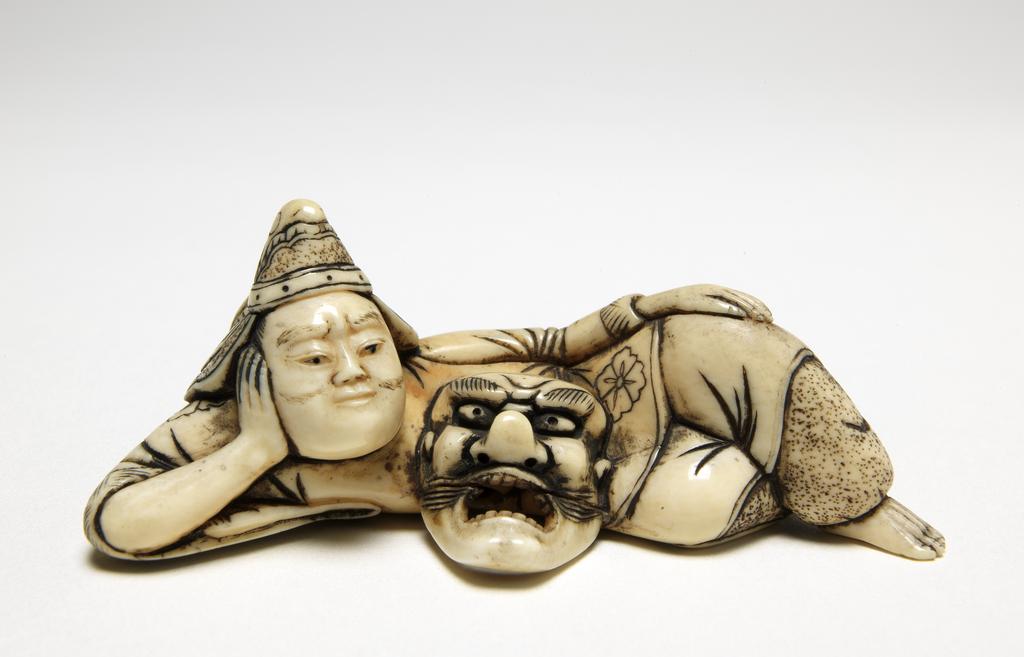 An image of Netsuke. A Bugaku dancer reclining, wearing his typical headgear and with his mask in front of him. Ivory, carved and stained, width 7 cm. Japanese.