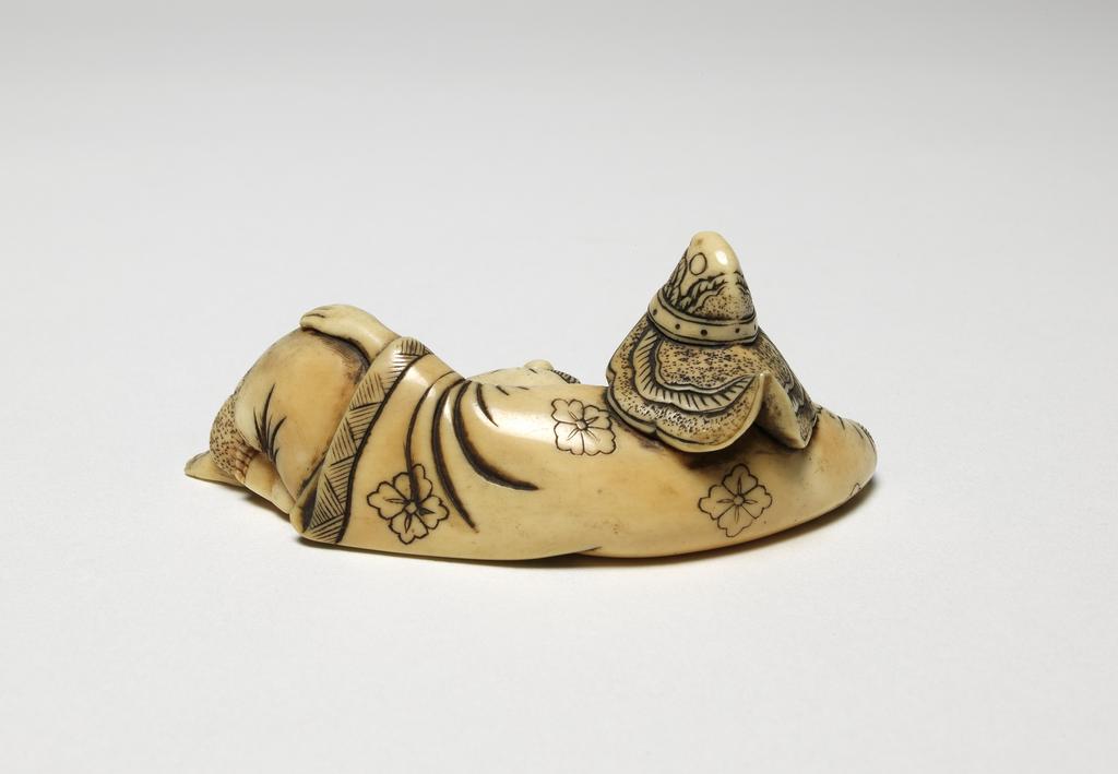 An image of Netsuke. A Bugaku dancer reclining, wearing his typical headgear and with his mask in front of him. Ivory, carved and stained, width 7 cm. Japanese.