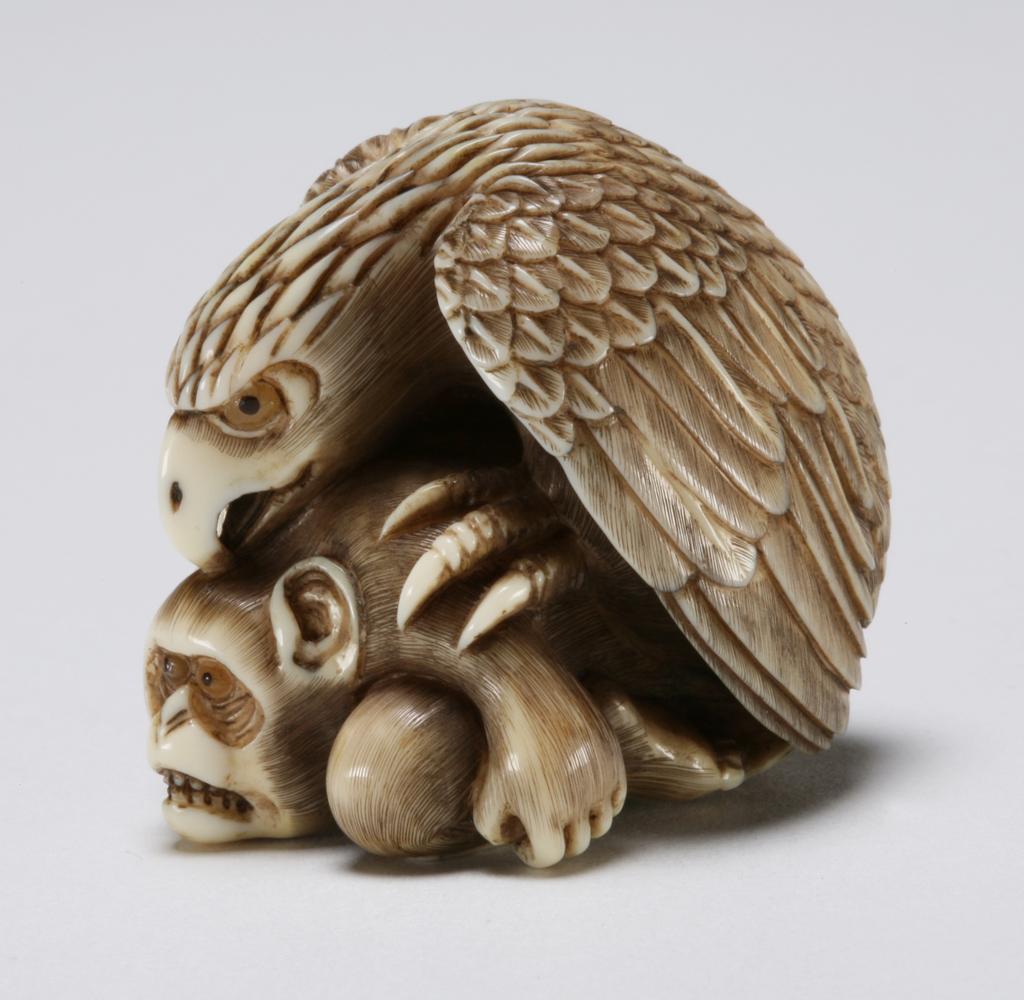 An image of Netsuke. A large eagle crouching and pecking the head of a monkey, its face is showing the fear. Ivory, carved, the eyes are double inlaid with glass, 1870-1930. Production Place: Japan. 