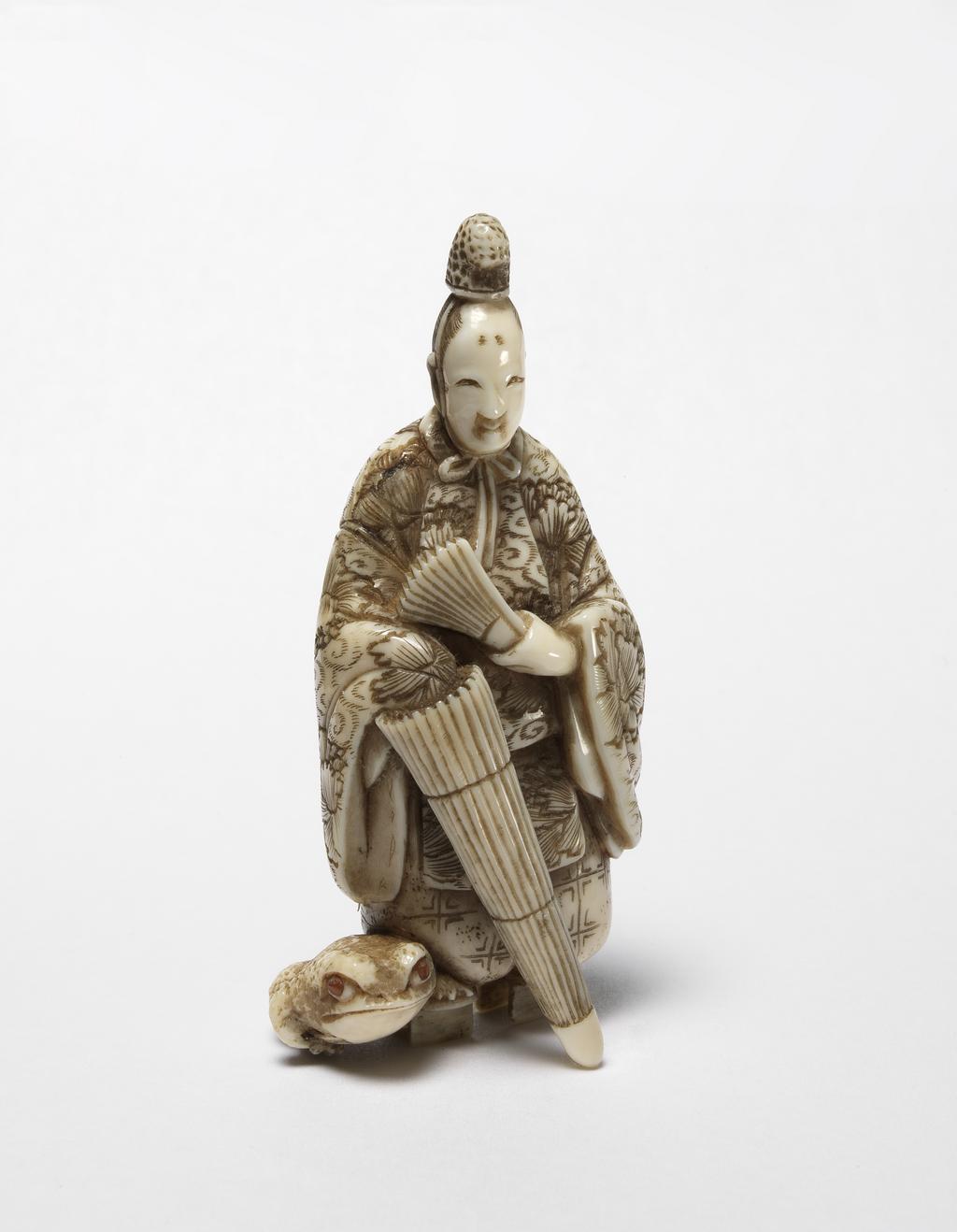 An image of Netsuke. Ono No Tofu in his ceremonial garments wearing Geta (wooden clogs) standing and holding his umbrella in his right hand and a folding fan in his left hand. A large frog is sitting next to his right foot. Ivory, carved, the eyes of frog are inlaid with horn, 1870-1900. Japanese.