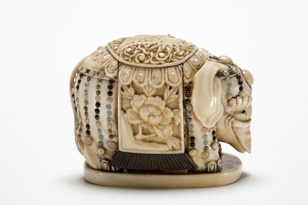 An image of Netsuke. Ivory and semi precious stones, an elephant of a Shibayama type, on a stand in full ceremonial decoration with a large  blanket beautifully embroidered of crane and peonies also chains of jewel strung from the blanket. Himotoshi ring in Mother of pearl. 1870-1900. Japanese.