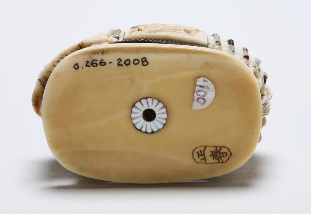 An image of Netsuke. Ivory and semi precious stones, an elephant of a Shibayama type, on a stand in full ceremonial decoration with a large  blanket beautifully embroidered of crane and peonies also chains of jewel strung from the blanket. Himotoshi ring in Mother of pearl. 1870-1900. Japanese.