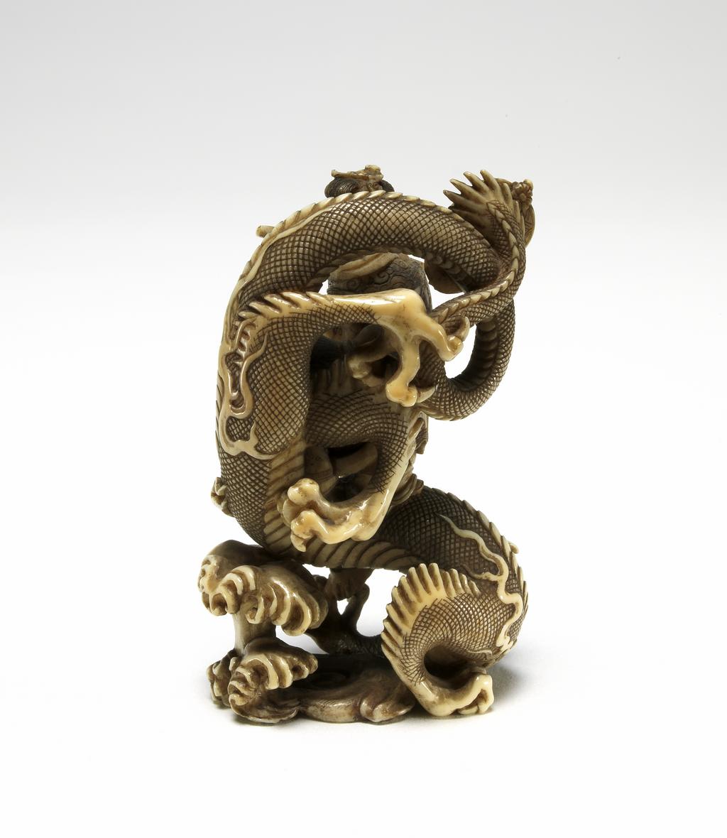 An image of Netsuke. Seiōbo, Queen Mother of the West, crowned with a dove and wearing a cloud and dragon patterned robe. Gyokushu (Japanese). Her attendant holds a fan and they are both standing on a three clawed dragon amongst waves. Middle of fan pole broken. Ivory, height 5.5 cm, width 3.5 cm, c. 1950. Japanese.