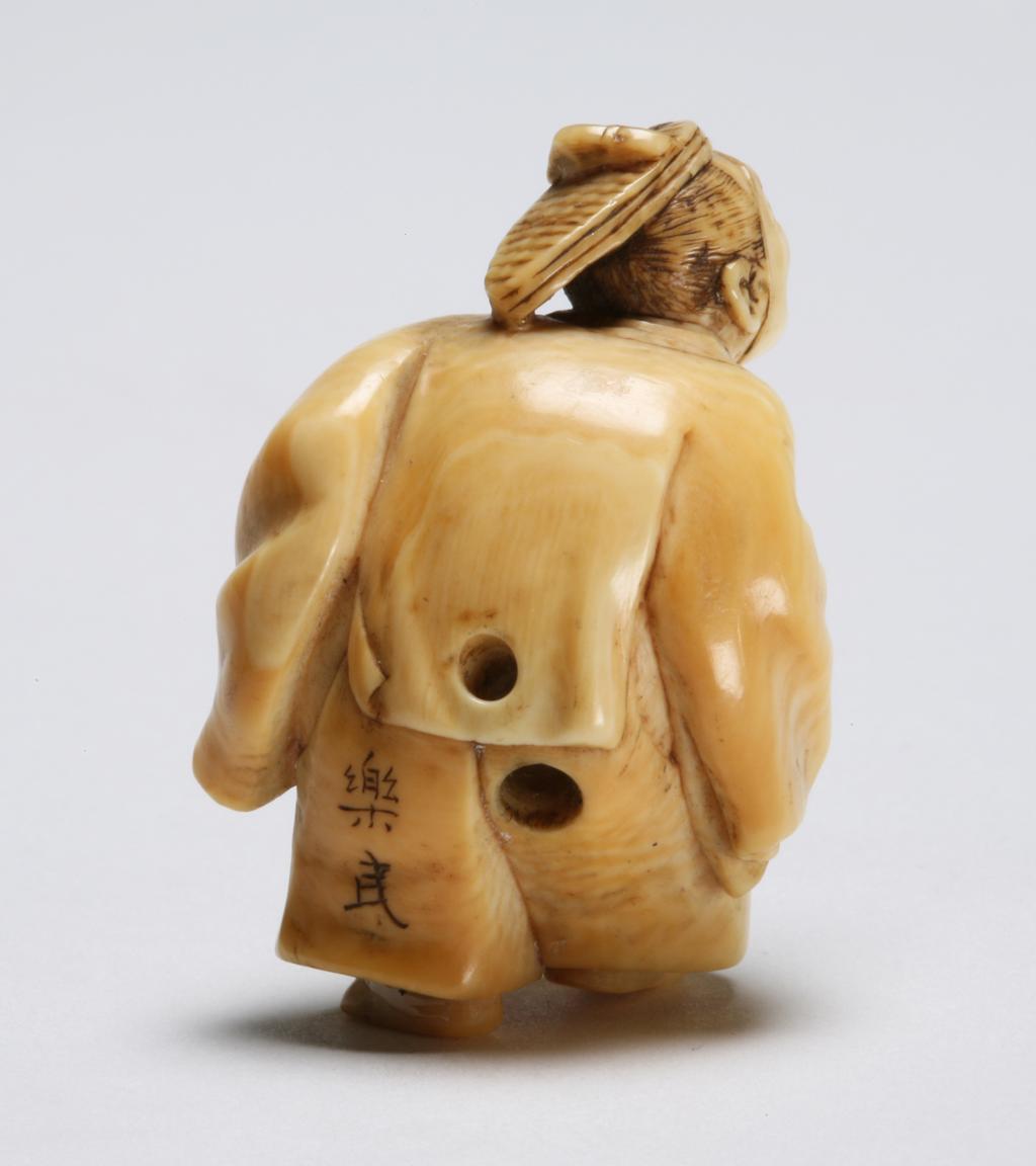 An image of Netsuke/katabori. Dancer. Rakumin, Edo Period (1603-1868). Smiling figure of a dancer wearing a kimono and a hat tied beneath his chin and holding a drum under his left arm. His hair and features are stained for effect. Himotoshi at the back. Ivory, carved and stained, circa 1800-1868. Edo Period.