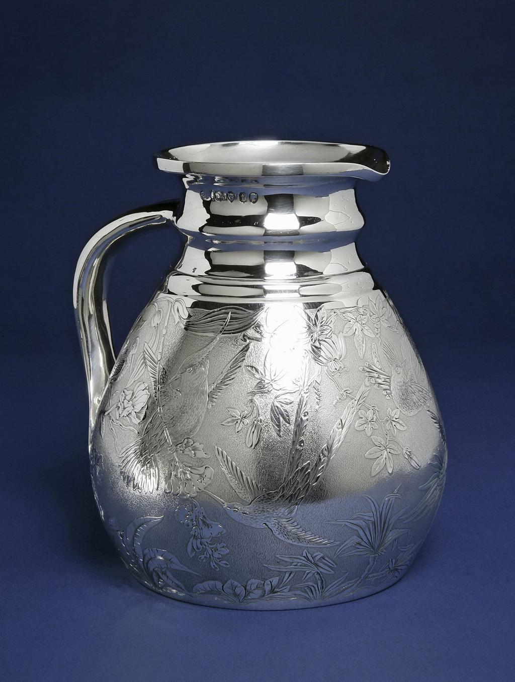 An image of Cambridge ale jug/water jug. Smith, Stephen (British). In the shape of a Cambridge ale jug, the body with sloping cylindrical sides and slightly tucked-in base and flat bottom. The narrow neck with plain moulded rings below a slightly everted rim. The broad handle is concave down the centre and joins the body near the base with a flat spatulate terminal. The sides of the body are matted and engraved with exotic birds and tropical flowers and foliage (including humming birds and passion flowers). The hollow handle was seamed down its length. The body was raised or spun, then the base neck and handle were soldered on. The body was engraved, and the interior gilt. Height, to the rim, 18.2 cm, width, overall, 17 cm, weight, whole, 845 g, 1874-1875. Aesthetic Movement. Victorian. England, London.