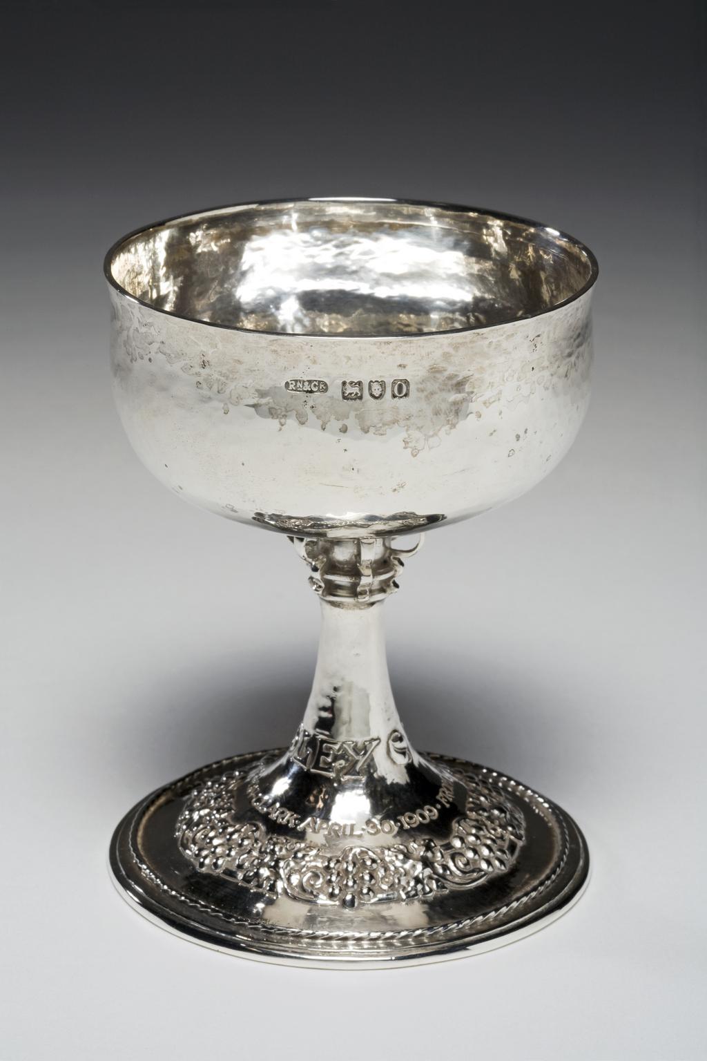 An image of Goblet. Ramsden & Carr. Ramsden, Omar (British, 1873-1939). Carr, Alwyn (British,1872-1940). Production Place:  England, London. The low circular bowl has bombé sides and a slightly everted rim. It is supported by a trumpet-shaped foot with six applied volutes at the top. The foot has an applied rope border set in from the edge, and is embossed and chased with a circle of fruiting vine. The lower part of the stem is embossed with ‘TO LESLEY’ and below is engraved ‘FROM HER GODFATHER C.S. GORDON CLARK APRIL 1909’. Silver, with hammered surface, embossed and chased decoration, and embossed, and engraved inscriptions. Height, overall, 10.6 cm, diameter, to rim, 8 cm, diameter, foot, 8.3, cm, weight, whole, 140 g, 1909. Arts and Crafts. Notes: It is unclear which silversmiths are associated with London.
