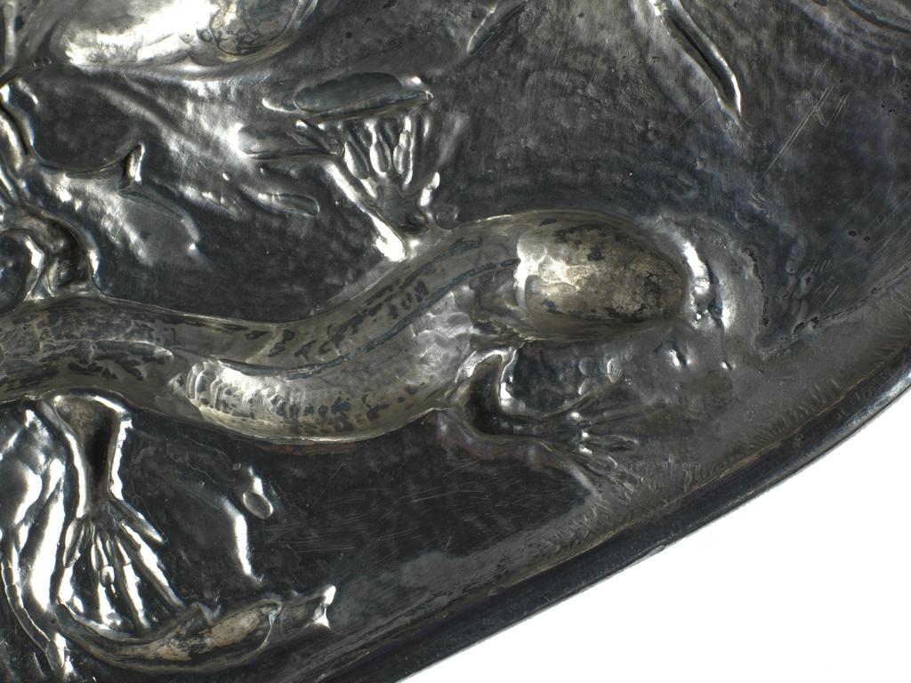 An image of Silver dish. Marks, Gilbert Leigh (British, 1861-1905). The dish was raised and the lizards and foliage embossed and chased. The lizards have a small piece of silver seamed under the jaw and neck, which allows the head to stand proud of the dish. The background to the dish is spot hammered. Height to rim 5 cm (variable), diameter (across rim) 42 cm, 1898-1899. Arts and Crafts movement.