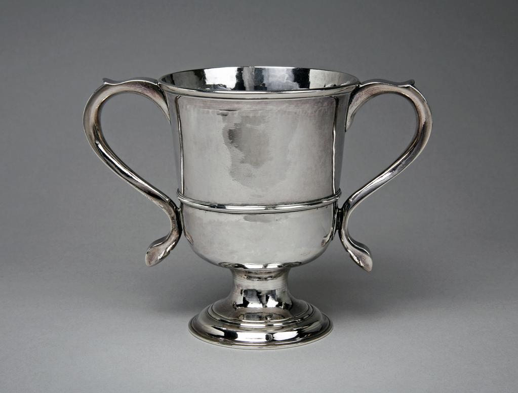An image of Two-handled loving cup. Langlands, John, I (British, free 1754; died 1793). The circular foot and stem raised in one piece, and supporting a deep cup with rounded base, straight sides, an applied horizontal moulding about a third of the way up, and a slightly everted rim. The plain S-scroll handles have a slight inward curve at the top to serve as a thumb rest. Near the top of one handle are the initials of a man and wife, 'M/I N'. Newcastle-upon-tyne, England. Silver, height, to rim, 13 cm, width, across handles, 18.1 cm, diameter, rim, 10.1 cm, c.1774-1775.
