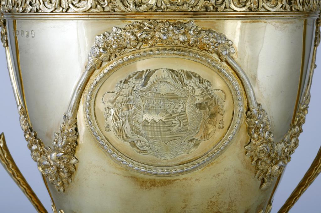 An image of Race Cup. Bateman, Peter. Bateman, Ann. Production Place:  London. The circular foot has a reeded edge above which is a band of leaf and bud ornament on a matted ground; the stem is embossed with bat's wing inverted fluting with an area of matting at the lower end of the flutes. The lower part of the tapering bowl has a calyx of lanceolate, wavy-edged leaves, above which, on each side of the body, is an oval medallion framed by husks. One medallion contains a plaque, cast and chased with two race horses and their jockeys racing in a landscape; the other is engraved with a coat of arms, sable a fess dancetty or in chief three fleur de lis argent impaling azure three uniorns' heads erased argent 2 and 1. Above each medallion there is an applied festoon of flowers and foliage from which depend two more festoons on either side, which loop up to meet the handles. The upper rim of the bowl has an applied frieze of strapwork and scrolling folliage within two moulded borders. The handles have lanceolate wavy-edged leaves at each upper end and a rib of husks on the exterior. The decoration of the cover follows that of the foot, but rises to a dome with reeded border topped by wavy edged leaves and an inverted pineapple finial. Decorated with cast and chased, applied, embossed and matted ornament and engraved with a coat of arms. Silver-gilt, height, overall, 59.9 cm, width, overall, 38.7 cm, 1799-1800. Neoclassical. Notes: The coat-of-arms on the dexter side are for Feeke, Feake or Feke of Wighton, Norfolk, or Hewster of Norfolk. The arms on the sinister side have not been identified, but could be for Shelley of Norfolk if the field was gules not azure.
