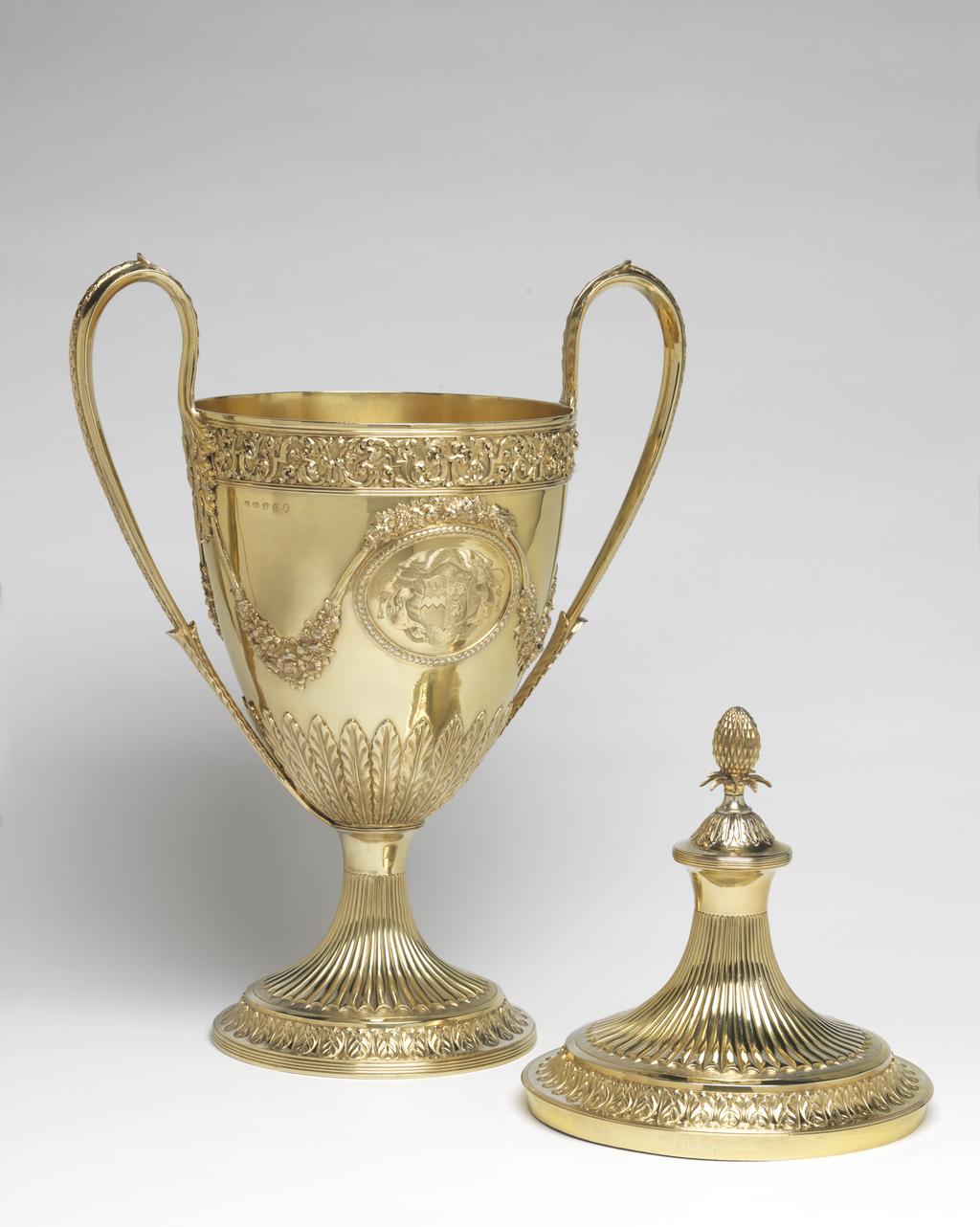 An image of Race Cup. Bateman, Peter. Bateman, Ann. Production Place:  London. The circular foot has a reeded edge above which is a band of leaf and bud ornament on a matted ground; the stem is embossed with bat's wing inverted fluting with an area of matting at the lower end of the flutes. The lower part of the tapering bowl has a calyx of lanceolate, wavy-edged leaves, above which, on each side of the body, is an oval medallion framed by husks. One medallion contains a plaque, cast and chased with two race horses and their jockeys racing in a landscape; the other is engraved with a coat of arms, sable a fess dancetty or in chief three fleur de lis argent impaling azure three uniorns' heads erased argent 2 and 1. Above each medallion there is an applied festoon of flowers and foliage from which depend two more festoons on either side, which loop up to meet the handles. The upper rim of the bowl has an applied frieze of strapwork and scrolling folliage within two moulded borders. The handles have lanceolate wavy-edged leaves at each upper end and a rib of husks on the exterior. The decoration of the cover follows that of the foot, but rises to a dome with reeded border topped by wavy edged leaves and an inverted pineapple finial. Decorated with cast and chased, applied, embossed and matted ornament and engraved with a coat of arms. Silver-gilt, height, overall, 59.9 cm, width, overall, 38.7 cm, 1799-1800. Neoclassical. Notes: The coat-of-arms on the dexter side are for Feeke, Feake or Feke of Wighton, Norfolk, or Hewster of Norfolk. The arms on the sinister side have not been identified, but could be for Shelley of Norfolk if the field was gules not azure.