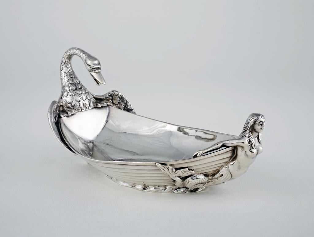 An image of Boat-shaped dish. Whitford, Samuel II, silversmith, England, London. Formed as a clinker-built ship on water, with a cast model of a mermaid at the prow and, at the stern, a cast model of a swan with neck arched to form a handle. Silver, the sides of the boat cast and the cast swan and mermaid applied, length, overall, 19 cm, weight, whole, 450 g, 1817-1818.