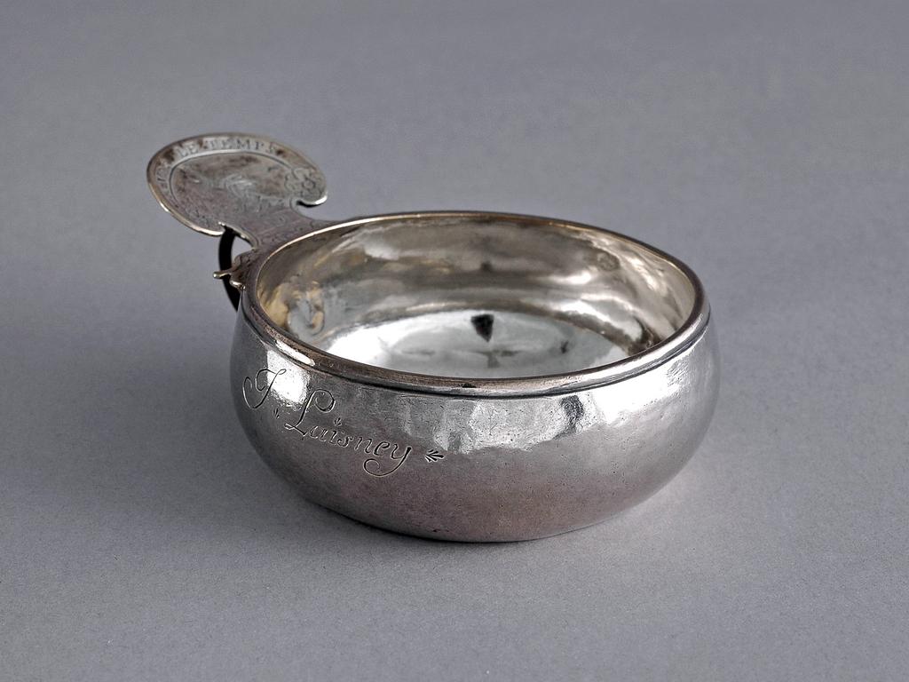 An image of Wine taster. Desnos, Pierre II, silversmith (French, 18th century). The circular bowl has slightly bulbous sides, and slightly raised base. The kidney shaped thumb-piece is attached to the bowl by leaf-shaped scrolls, undher wish is a finger ring. The upper surface is inscribed 'AVEC LE TEMPS' and engraved with a flaming heart and a cupid holding a hammer. The side of the bowl is inscribed 'J Laisney'. Silver, height, to top of handle, 4.4 cm, width, rim to handle, 11.5 cm, weight, whole, 120 g, 1754. Louis XV.