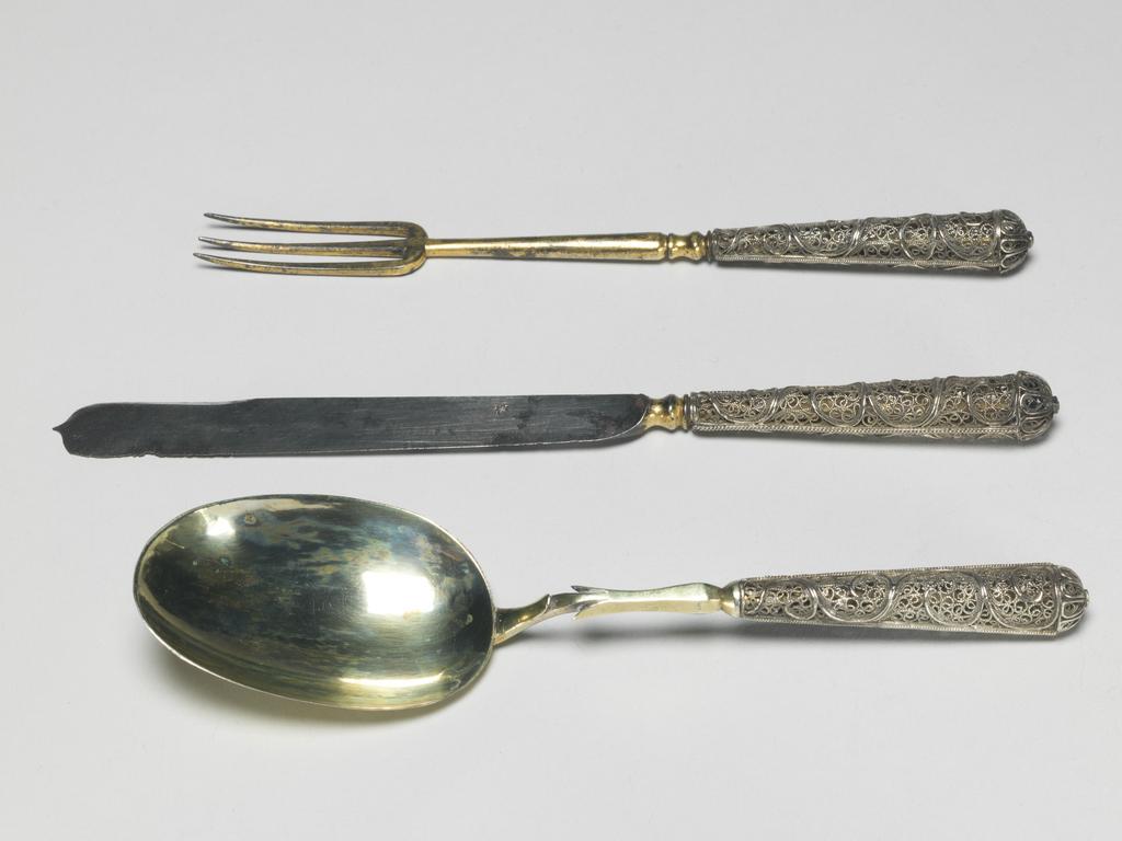 An image of Cutlery Set. Knife, fork, spoon and case. Laminet, Johann Christoph, spoon maker, attributed, Augsburg. Unidentified cutler. Unknown handle maker. Silver, parcel-gilt and steel, leather with gilt tooling; the knife (A) has a steel blade which flares towards its rounded end with central point; the baluster shaped bolster is gilt; the silver handle is circular in section and broadens towards the terminal cap; the sides are of open filigree scrolls between two rope borders. The fork (B) has three gilt metal tines on a long knopped stem. The spoon has an oval silver-gilt bowl with a rat-tail on the back. Both the fork and spoon have handles which are similar to that of the knife. The case (C) has a hinged lid and is approximately the shape of a spoon with a broad handle. It is covered with red leather with tooled partially gilt decoration. Length, knife, 17.7 cm, length, fork, 15.4 cm, length, spoon, 15.7 cm, length, case, 19.3 cm, circa 1695- circa 1707. German.