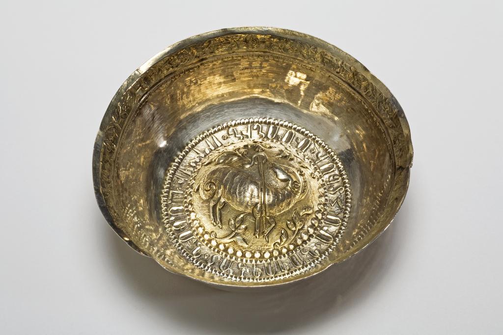 An image of Bowl. Wine Cup/Wine Taster. Unknown silversmith, Balkans, possibly. No hallmarks or maker's marks. The bowl has an everted rim and domed base. The rim is gilt and chased with a border of stylised flowers on a matted ground, beneath is a punched border. The base is gilt and embossed with an Agnus Dei; the stylised lamb on a matted ground embossed with leaves and a border of beads, and surrounded by a Cyrillic inscription. Silver, parcel-gilt, raised, embossed, chased, matted and punched, height, to rim, 3.5 cm, approximate, diameter, across rim, 11.3 cm, approximate, weight, whole, 62 g.