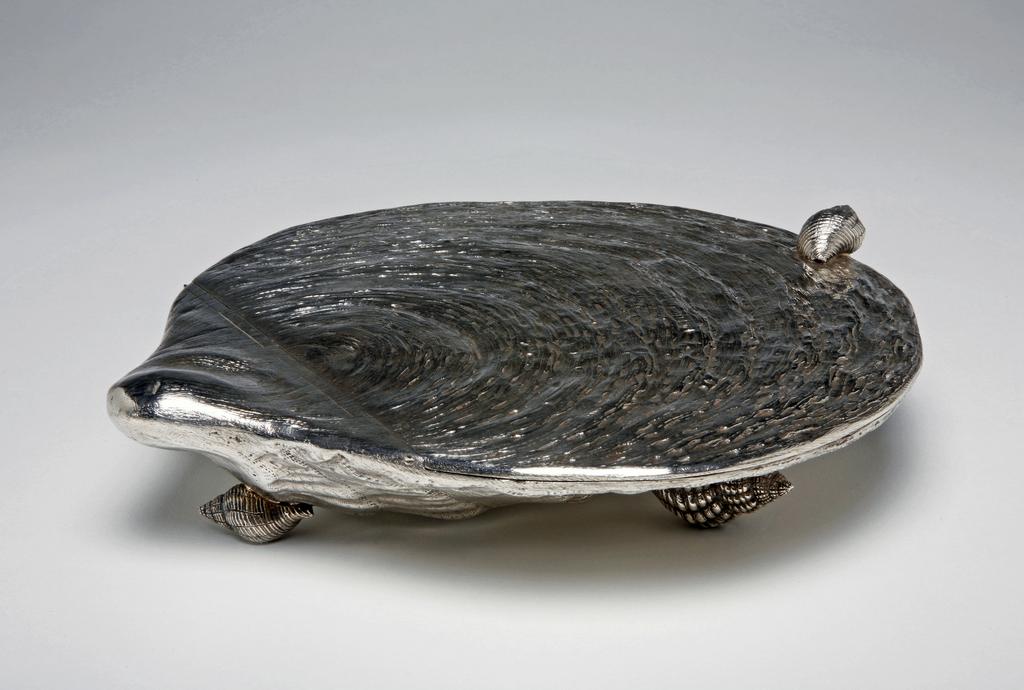 An image of Shell-shaped box. Angel, George, England, London. The cast escallop shell-shaped box has a flat cover with concealed hinge, and a cast winkle shell-shaped handle, and stands on two large and one small cast winkle shell-shaped feet. The whole is realistically chased. The cast body seamed with the back of the cover and set with a hinge. The edge of the cover and body has an applied rim mount. The handle and feet are cast and applied. Silver, height, to top of handle, 5.3 cm, width, handle to back, 22.8 cm, weight, whole, 640 g, 1875-1876. Victorian.