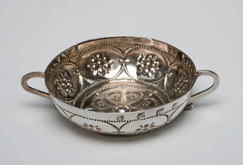 An image of Silver wine tasterSilver; shallow bowl between two flattened scroll handles; the sides punched with beaded borders and eight oval cartouches containing embossed flowerheads; the base punched and embossed with an eight petaled flower;  initialled 'WHWI' followed by a leafC.1646-7London, England