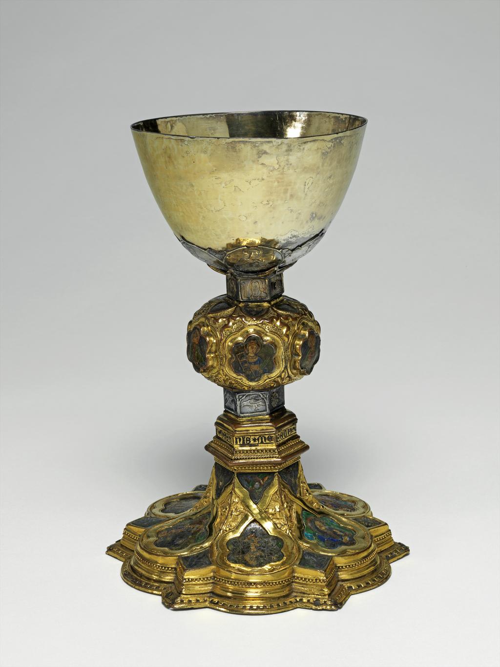 An image of Silver/Church Plate. Chalice. Petruccio Campagnini, Andrea, probably (active Tuscany, Siena, Italy). Set with six silver medallions of Christ, St Peter, St. Paul, St. John, St. Bartholomew and possibly St. Barnabus. Above the knop the stem is engraved with six male busts without nimbi; below the knop the stem is engraved with six birds. At the base of the stem are moulded borders containing a plain band engraved and enamelled with the inscription 'ANDREA * PETRUCI * DE * SENIS * ME *FECI(T). The low copper-gilt trumpet-shaped foot has a shaped circular spreading base with rim of twelve alternating semicicular and pointed lobes. It is set with six applied silver plaques with translucent enamels on sunk relief, representing the Crucifixion with Instruments of the Passion, on either side of which are the Virgin and St. John; on the opposite side are a bishop with mitre and crozier between a male and female saint. Above are smaller shaped triangular plaques enamelled with the heads of angels; and below are lozenge shaped plaques enamelled with a stylised leaf. The base stands on a foot ring decorated with moulded, beaded and dentil borders. Bowl silver-gilt with silver calyx, foot and stem of copper-gilt, decorated with embossing and chasing, and set with silver plaques with basse taille enamelling, height, overall, 19 cm, diameter, base, 12.8 cm, maximum, diameter, rim, 9.9 cm, circa 1370. Late Medieval. Gothic.