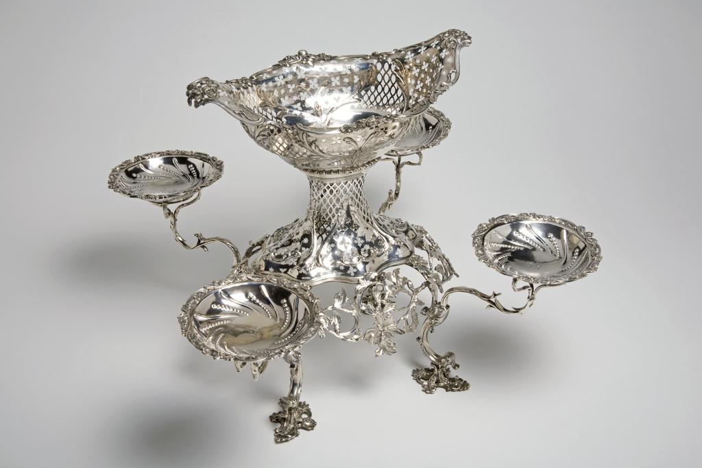An image of Epergne/Centre-Piece. Romer, Emick (Norwegian goldsmith, silversmith, 1724 to circa 1795, London). The oval openwork stand holding a central basket, with four brackets holding four small baskets. The top of the stand has a gadroon border above spreading sides, which are pierced with panels of quatrefoils, stars and lozenges between embossed scrolls. There are four cast double-scrolled legs suspended with cast and chased scrolling foliage, standing on four ornate openwork cast rocaille feet. The detachable lozenge-shaped basket is similarly pierced and embossed, with an applied openwork border of scrolls and flowers. The four scrolling branches have applied leaves. They attach to the top of the legs, and support four low circular dishes embossed with scrolling flutes and beads within an openwork applied border of scrolls and flowers. Each of the bowls is engraved with a crest of a greyhound courant in front of an oak tree for Daly, Kennedy, Molloy, or O'Mulloy. The stand and baskets are raised and pierced with applied decoration. The legs and branches are cast with applied decoration. Silver, height, overall, 35 cm, width, central basket, 28 cm, weight, whole, 2260 g, circa 1759-1760. English. Rococo.