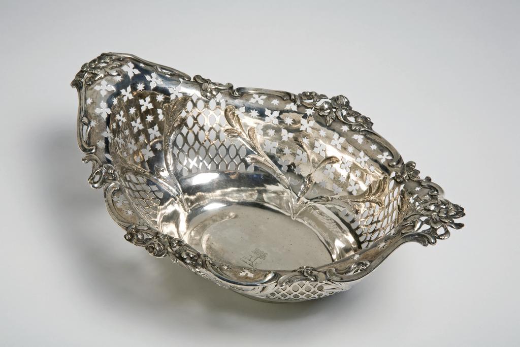 An image of Epergne/Centre-Piece. Romer, Emick (Norwegian goldsmith, silversmith, 1724 to circa 1795, London). The oval openwork stand holding a central basket, with four brackets holding four small baskets. The top of the stand has a gadroon border above spreading sides, which are pierced with panels of quatrefoils, stars and lozenges between embossed scrolls. There are four cast double-scrolled legs suspended with cast and chased scrolling foliage, standing on four ornate openwork cast rocaille feet. The detachable lozenge-shaped basket is similarly pierced and embossed, with an applied openwork border of scrolls and flowers. The four scrolling branches have applied leaves. They attach to the top of the legs, and support four low circular dishes embossed with scrolling flutes and beads within an openwork applied border of scrolls and flowers. Each of the bowls is engraved with a crest of a greyhound courant in front of an oak tree for Daly, Kennedy, Molloy, or O'Mulloy. The stand and baskets are raised and pierced with applied decoration. The legs and branches are cast with applied decoration. Silver, height, overall, 35 cm, width, central basket, 28 cm, weight, whole, 2260 g, circa 1759-1760. English. Rococo.