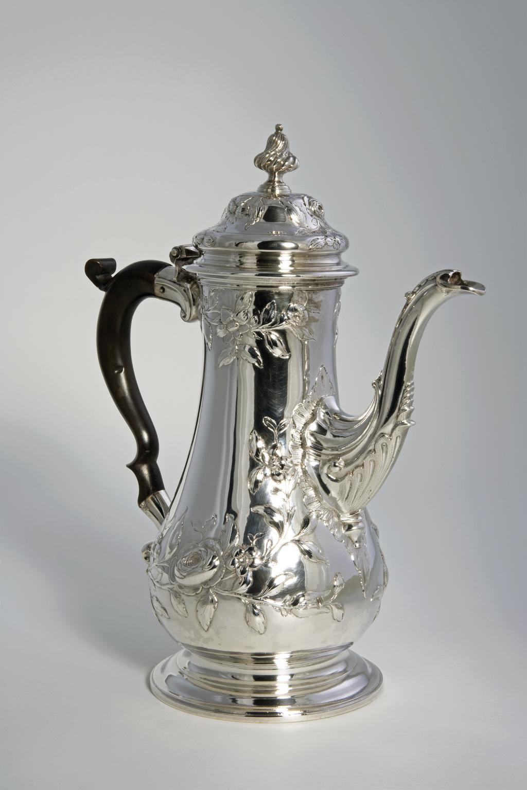 An image of Coffee Pot. Swift, John (British). The tall pear-shaped body is embossed with sprays of leaves and flowers. The top of the swan's neck spout is leaf capped, the lower part embossed with flutes and a stylised shell. The body stands on a spreading circular foot. The domed cover is embossed with sprays of flowers and has a wythern silver knop. The double scroll handle is of wood. Silver, embossed and chased, height, to knop, 28.8 cm, width, spout to handle, 23.5 cm, weight, gross, 1020 g, circa 1763-1764. Production Place: London, England.  Rococo.