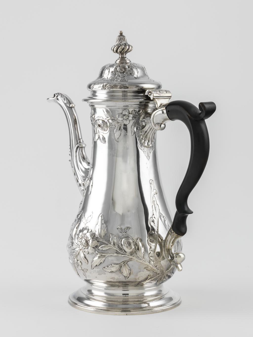 An image of Coffee Pot. Swift, John (British). The tall pear-shaped body is embossed with sprays of leaves and flowers. The top of the swan's neck spout is leaf capped, the lower part embossed with flutes and a stylised shell. The body stands on a spreading circular foot. The domed cover is embossed with sprays of flowers and has a wythern silver knop. The double scroll handle is of wood. Silver, embossed and chased, height, to knop, 28.8 cm, width, spout to handle, 23.5 cm, weight, gross, 1020 g, circa 1763-1764. Production Place: London, England.  Rococo.