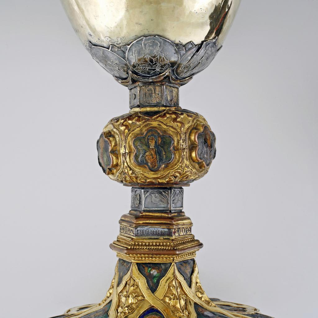 An image of Silver/Church Plate. Chalice. Petruccio Campagnini, Andrea, probably (active Tuscany, Siena, Italy). Set with six silver medallions of Christ, St Peter, St. Paul, St. John, St. Bartholomew and possibly St. Barnabus. Above the knop the stem is engraved with six male busts without nimbi; below the knop the stem is engraved with six birds. At the base of the stem are moulded borders containing a plain band engraved and enamelled with the inscription 'ANDREA * PETRUCI * DE * SENIS * ME *FECI(T). The low copper-gilt trumpet-shaped foot has a shaped circular spreading base with rim of twelve alternating semicicular and pointed lobes. It is set with six applied silver plaques with translucent enamels on sunk relief, representing the Crucifixion with Instruments of the Passion, on either side of which are the Virgin and St. John; on the opposite side are a bishop with mitre and crozier between a male and female saint. Above are smaller shaped triangular plaques enamelled with the heads of angels; and below are lozenge shaped plaques enamelled with a stylised leaf. The base stands on a foot ring decorated with moulded, beaded and dentil borders. Bowl silver-gilt with silver calyx, foot and stem of copper-gilt, decorated with embossing and chasing, and set with silver plaques with basse taille enamelling, height, overall, 19 cm, diameter, base, 12.8 cm, maximum, diameter, rim, 9.9 cm, circa 1370. Late Medieval. Gothic.
