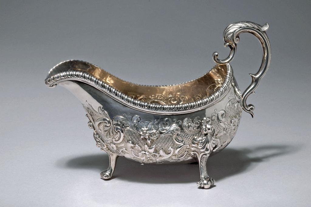 An image of Sauce boat. W?, silversmith, Ireland, Dublin. The large, deep oval bowl has a gadroon edged border; the sides are embossed and chased with scrolls, leaves, flowers, matted diaperwork, scalework and a vacant cartouche on one side (later decorated). It stands on three cast and chased scroll legs with ornate shell capitals and scroll feet and has a chased leaf-capped triple scroll handle. Silver, height, to top of handle, 15.5 cm, width, from lip to handle, 22.5 cm, weight, overall, 560 g, circa 1750. Rococo.